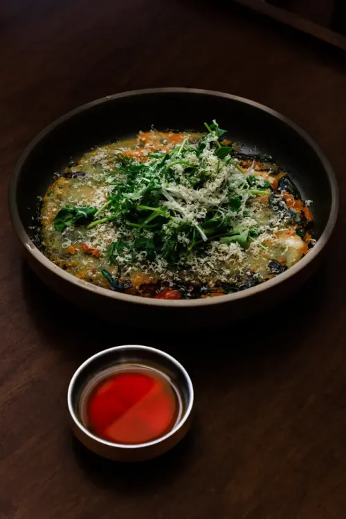 A savoury pancake topped with herbs and grated cheese, with a dipping sauce