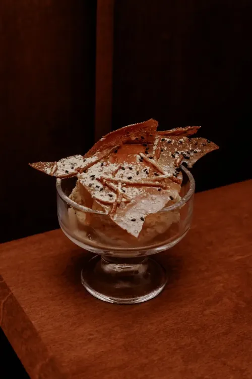 A dessert in a glass cup topped with sugar-dusted crisps and black sesame