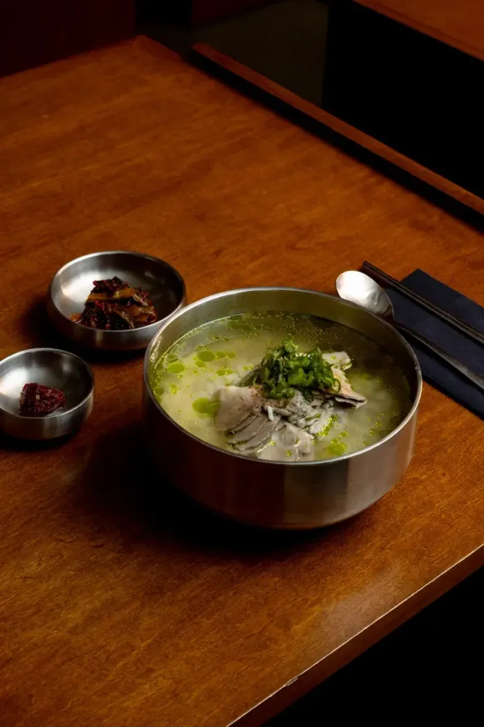 A steel bowl of gomtang with two small side dishes on a wooden table