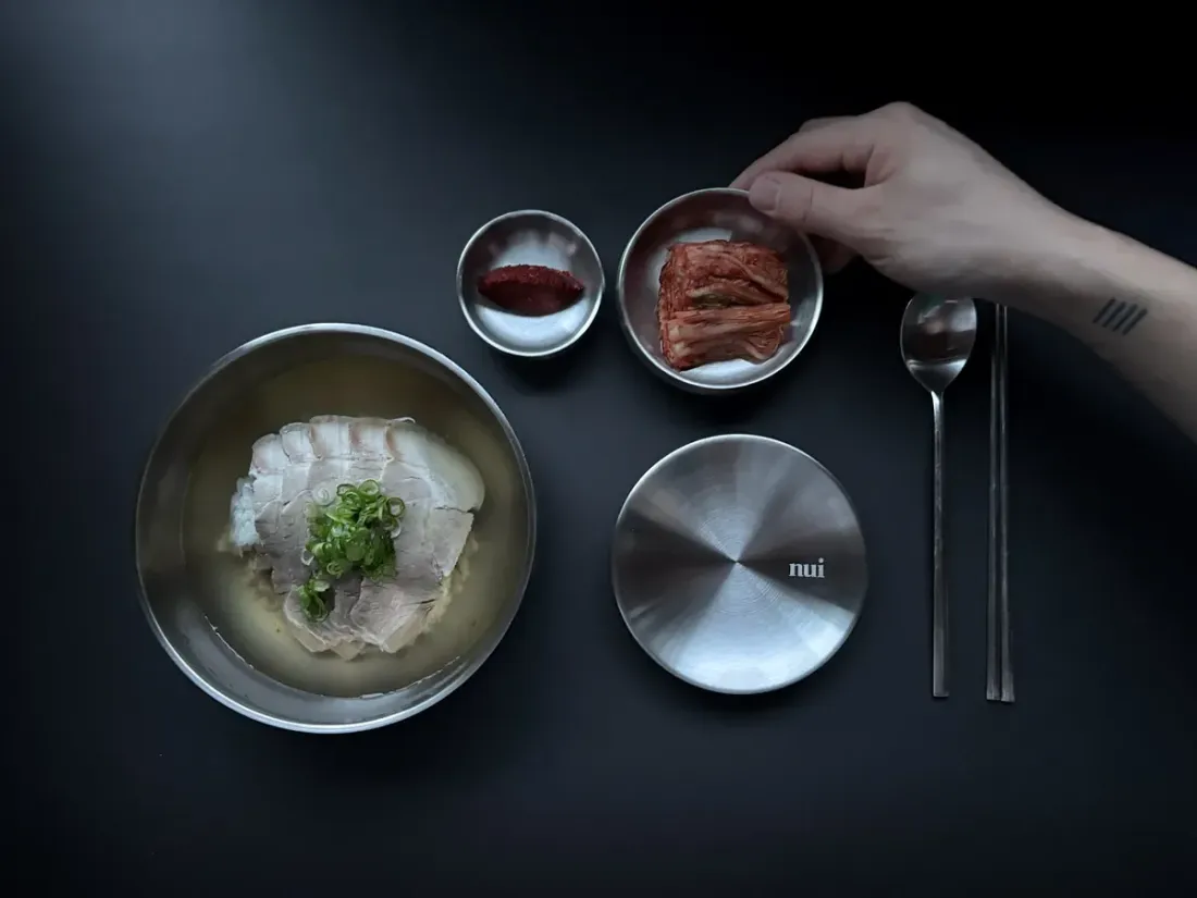 A table set from above with gomtang, kimchi, a Nui bowl lid and cutlery